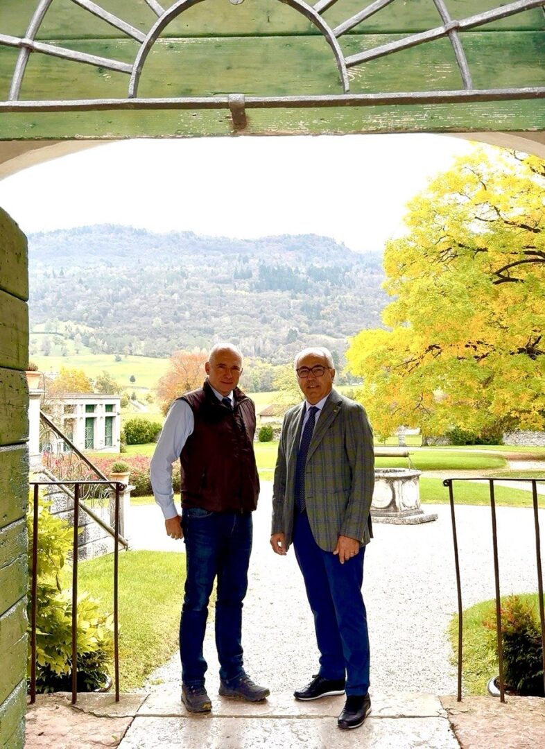 Foto che raffigura Presidente Pozza e Conte Fulcis in piedi con lo scorcio delle montagne all'interno del giardino della VIlla di Modolo