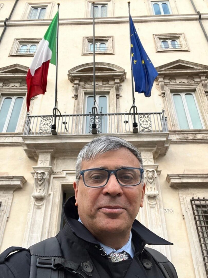 Foto mezzo busto di Anilkumar Dave - Space Economy advisor con sfondo di un palazzo istituzionale con affisse le bandiere italiana e europea